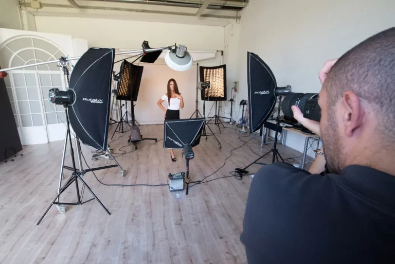 Fotografo professionista scatta foto in uno studio ben attrezzato, con una modella in posa di fronte a diversi softbox e luci di studio. L'ambiente è spazioso e luminoso, con pavimento in legno e pareti bianche.