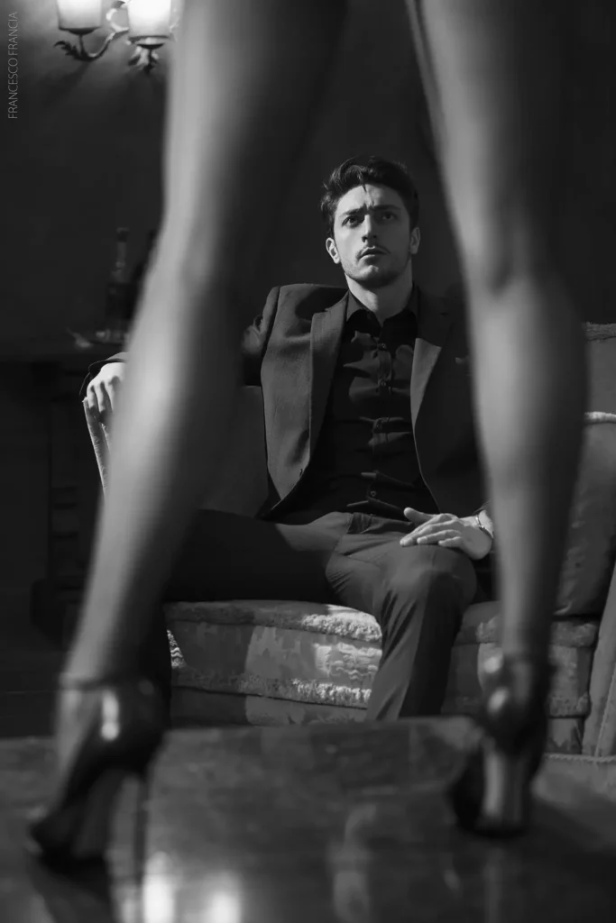 Uomo elegante in abito scuro osserva una figura femminile in tacchi alti in un'ambientazione raffinata