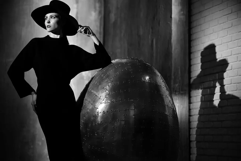 fotografia di moda di Francesco Francia per una campagna pubblicitaria di abiti Cashmere. Modella con abito nero e cappello poggiata su un grande uovo metallico illuminata da una luce drammatica stile Helmut newthon con ombre dure e luci cinematografiche